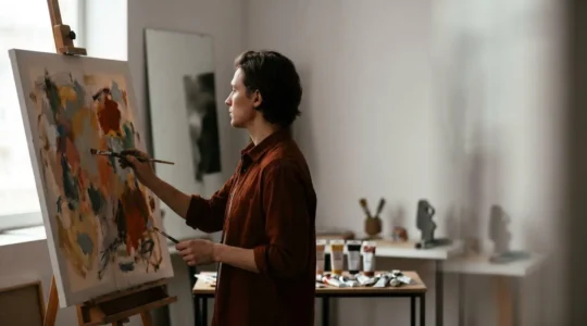 An artist contemplating their reflection while creating a traditional self-portrait in a sunlit studio